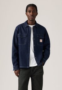 Chemise en denim bleu foncé, boutonnée à l'avant, deux poches poitrine, étiquette blanche avec logo, portée sur un t-shirt blanc, associée à un pantalon foncé.