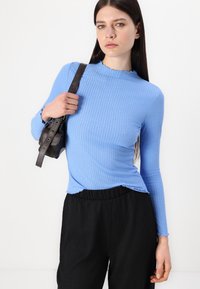 Top a maniche lunghe ribattezzato blu chiaro con scollo a ondine, abbinato a pantaloni neri textured e in mano una piccola borsa marrone.