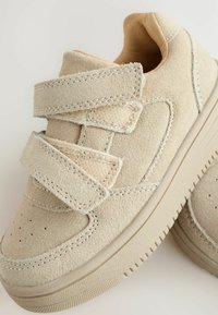 Baskets en suède beige avec deux sangles Velcro et une semelle texturée, posées sur un fond clair.