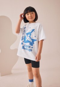 T-shirt en coton blanc avec un motif graphique bleu représentant des étoiles et du texte. Porté avec un short noir et des baskets montantes blanches avec des accents bleus.