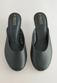 Schwarze Slip-On-Schuhe aus glattem synthetischem Material, mit goldenen Nieten entlang der Außensohle und einem abgerundeten Zehendesign.