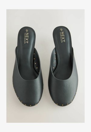 Pantofi slip-on negri realizați din material sintetic neted, cu nituri aurii de-a lungul talpii și cu design rotund al vârfului.