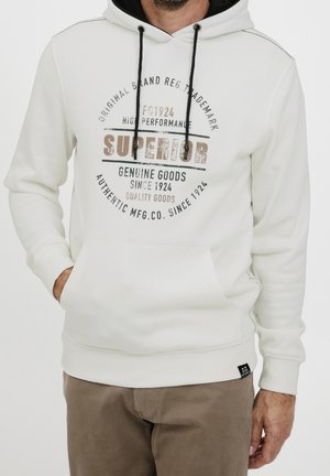 Mężczyzna noszący białą bluzę z kapturem z czarnymi sznurkami i brązowym napisem "Superior Genuine Goods Since 1924,", w połączeniu z brązowymi spodniami.
