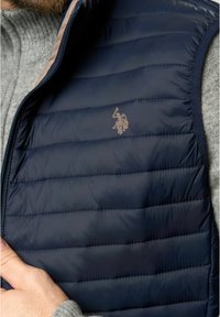 Gilet imbottito navy con un design trapuntato, rifinito con un bordo beige e un piccolo logo in rilievo. Texture morbida, chiusura frontale con zip.