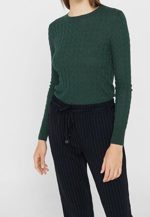 Personne portant un haut texturé vert foncé à manches longues et un pantalon noir à fines rayures avec une ceinture nouée, debout devant un fond uni.