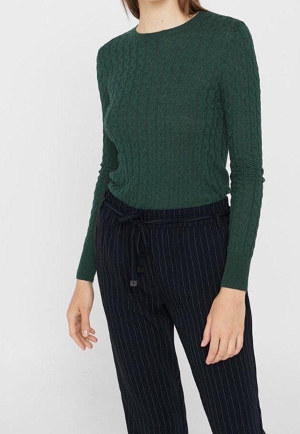Personne portant un haut texturé vert foncé à manches longues et un pantalon noir à fines rayures avec une ceinture nouée, debout devant un fond uni.
