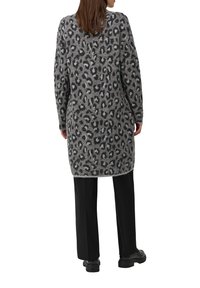 Femme debout, portant un long cardigan gris à imprimé léopard, un pantalon noir et des chaussures noires, vue de dos sur un fond blanc.