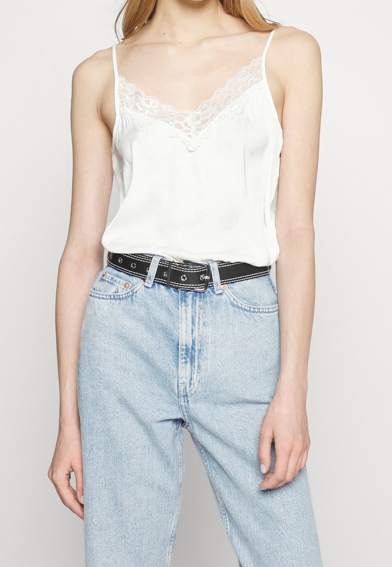 Femme portant un caraco blanc bordé de dentelle, rentré dans un jean bleu clair taille haute avec une ceinture noire sur un fond uni.