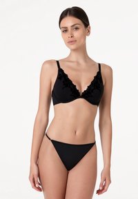 Donna che indossa un completo intimo nero con dettagli in pizzo floreale sulle coppe del reggiseno, su uno sfondo bianco semplice.