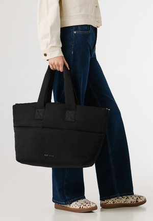 Persoon die donkerblauwe jeans en een beige shirt draagt, houdt een grote zwarte Pepe Jeans boodschappentas vast en draagt witte schoenen met patroon.