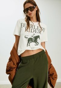 Hvid cropped t-shirt med "Fearless Riders" grafik, parret med olivengrønne joggere med sidelister, og en brun jakke draperet over skuldrerne.