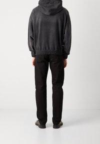 Schwarzer verkürzter Kapuzenpullover mit Zugbandkapuze und elastischem Bund, kombiniert mit schwarzen geraden Hosen und robusten Schuhen, von hinten betrachtet.
