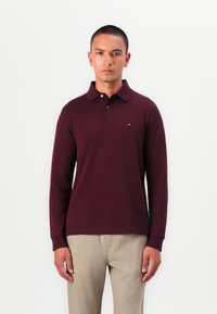 Maroon long-sleeve polo shirt with a collar, two-button placket, and a small logo. Worn with light beige pants. Smooth fabric texture.