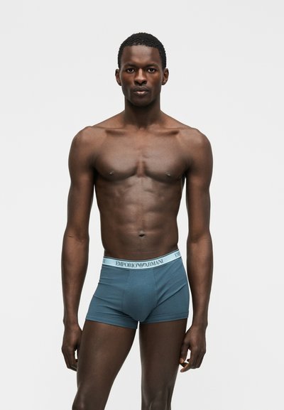 Αθλητικός άνδρας με σκουρόχρωμο δέρμα που φορά μπλε αντρικά μποξεράκια Emporio Armani, στέκεται με το πρόσωπο προς τα εμπρός μπροστά σε απλό ανοιχτό φόντο.