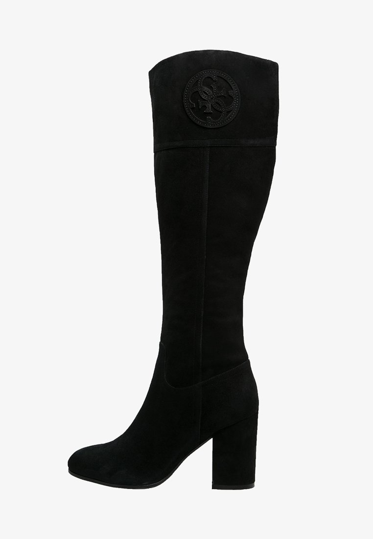 Botas de ante negras por encima de la rodilla con un tacón cuadrado, que presentan un emblema circular en la parte superior y una textura de superficie lisa.