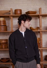 Črni rebrasti pleteni kardigan z okrolim izrezom, gumbi spredaj in stranskimi žepi. Postavljen pred lesene sklede in police v ozadju.