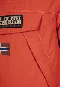 Oranssi ulkovaatetus, jossa on suuri logopatchi, edessä ommeltu tasku ja suorakulmainen kangaspäällyste. Sileä, teksturoitu materiaali.