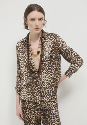 Donna che indossa una audace blusa con stampa leopardata e pantaloni abbinati, con due collane di perline a strati, in piedi con una mano sulla vita.