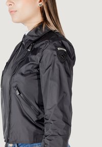 Bomber jacket leggero nero con tasche con zip, un'etichetta con logo sulla manica e un design aderente. Texture liscia e materiale idrorepellente.