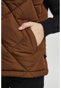 Un gilet matelassé marron avec un poignet de sweater côtelé noir contrastant. Il possède une poche latérale avec une finition lisse et un motif de couture diagonal.