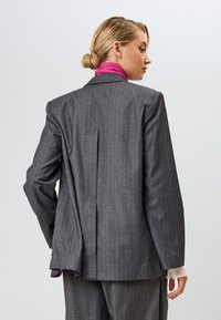 Harmaan raidoitettu bleiseri, jossa on kontrastivärinen vaaleanpunainen kaulus, takana tuuletusaukko ja hihoista pilkistävät teksturoidut pitsikämmenet.