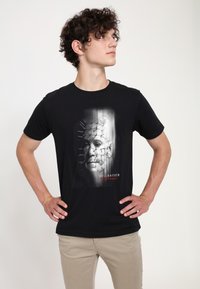 Svart bomullströja med en grafisk tryck av ett ansikte med spikar, kallad "Hellraiser," i vit och röd text. Kort ärm och design med rund hals.