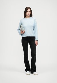 Lyseblå genser med lange ermer kombinert med svarte vide jeans. Personen holder en grå stoffgjenstand og står foran en nøytral bakgrunn.