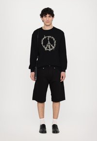 Black ribbed sweater featuring a peace symbol design, paired with black shorts. Model wears black loafers and white socks, holding a small bag.