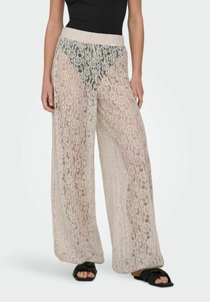 Pantalon beige à jambes larges en dentelle transparente porté sur des sous-vêtements noirs, associé à des sandales plates noires ouvertes à l'avant et un haut noir sans manches.