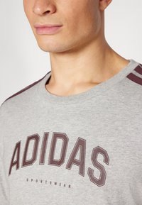 Pelēks kokvilnas T-krekls ar lielu tumši bordo "ADIDAS" logotipu, apaļu kakla izgriezumu un bordo svītrām uz pleciem. Klasisks piegriezums.