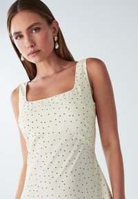 Cremefarbenes Kleid mit quadratischem Ausschnitt, versehen mit schwarzen Punkten, dünnen Trägern und einer leicht ausgestellten Silhouette. Glatter, leichter Stoff.