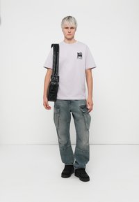 Lys lilla t-shirt med grafik og tekst, parret med falmede blå cargo-bukser med flere lommer, båret sammen med en sort, oversized skuldertaske.