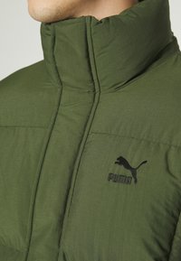 Veste matelassée vert olive avec col montant et logo Puma noir brodé. Présente une texture matelassée et une surface lisse.