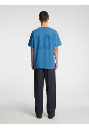 Personne debout de dos portant un t-shirt bleu avec le numéro 10 dans le dos et un pantalon noir ample, sur un fond blanc uni.