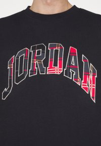 Sudadera de algodón negra con la palabra "JORDAN" en letras de cuadros bordadas, con acentos en rojo, gris y beige. Mangas cortas y cuello redondo.