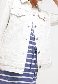 Veste en denim blanc avec des fermetures à boutons, deux poches poitrine et un ourlet effiloché. Portée par-dessus une robe rayée bleu et blanc avec un cordon de serrage.
