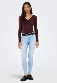 Μακρυμάνικη μπλούζα βερμούδα μπορντό με λεπτομέρεια σούρας στο V-λαιμό, συνδυασμένη με ανοιχτό μπλε στενά τζιν και μαύρα μοκασίνια, με μαύρη ζώνη.