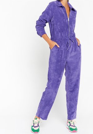 Persona che indossa una tuta in velluto a coste viola con zip frontale e vita regolabile, mani nelle tasche e sneaker colorate verde, viola e bianche.