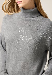 Maglione grigio a collo alto realizzato in tessuto a maglia, con un motivo decorativo e ornamenti lucenti sulla parte anteriore. Vestibilità comoda.