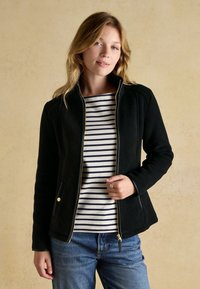 Frau mit hellen welligen Haaren, die eine schwarze Reißverschlussjacke über einem weißen Shirt mit schwarzen horizontalen Streifen und blauen Jeans trägt, vor einem beigen Hintergrund stehend.