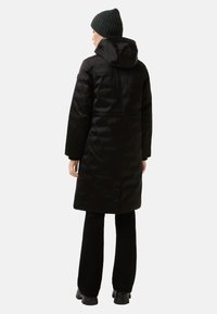 Zwarte geïsoleerde jas met een capuchon, voorzien van een rits aan de voorkant, zijzakken en een knoopsluiting aan de achterkant. Gecombineerd met een donkergroene muts.