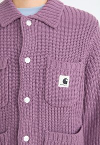 Lila gestrickte Strickjacke mit einem strukturierten Rippmuster, zwei vorderen Taschen, weißen Knöpfen und einem Carhartt-Logo-Patch.