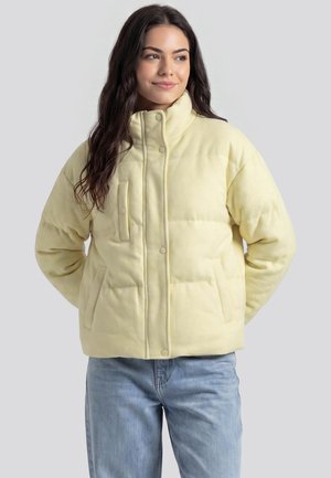 Junge Frau mit langen dunklen Haaren, die eine hellgelbe Daunenjacke und hellblaue Jeans trägt, lächelt und schaut zur Seite.
