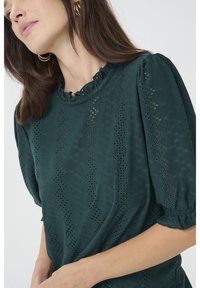 Blusa teal con un motivo testurizzato, caratterizzata da maniche a sbuffo e un collo arricciato. Il tessuto presenta dettagli a taglio in tutto il capo.