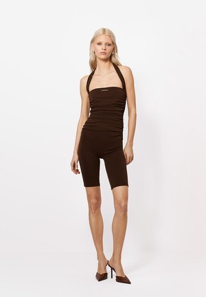 Szőke nő ujjatlan barna halter nyakú kezeslábasban és barna magassarkú cipőben áll fehér háttér előtt.