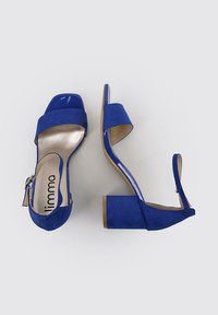 Tacones abiertos de ante azul con una correa brillante, un tacón cuadrado y una cremallera en la parte trasera. Cuenta con una plantilla beige con la marca.