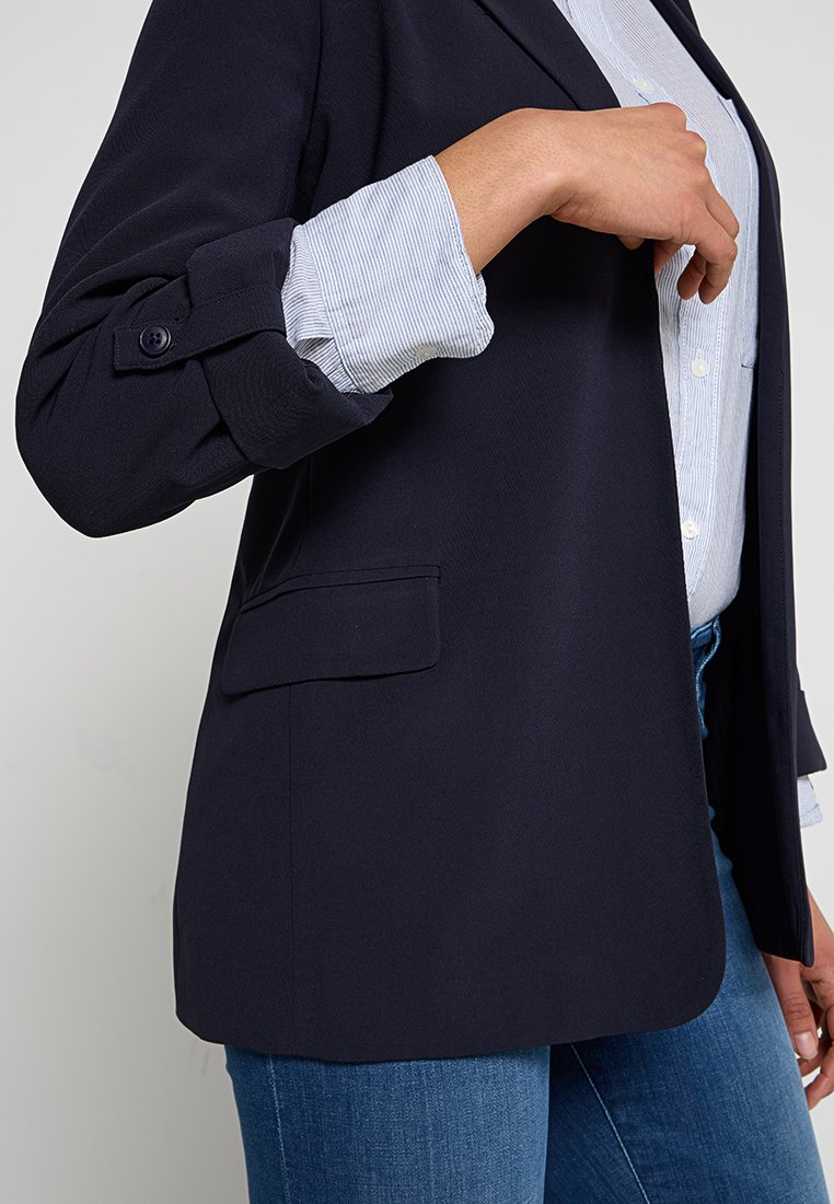 Blazer navy con un design strutturato e una texture liscia. Presenta una tasca singola, polsini con bottoni e è abbinato a una camicia a righe sotto.