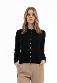 Cardigan nero in tessuto morbido, con scollatura tonda, maniche lunghe e bottoni dorati sul davanti. Indossato con pantaloni beige.