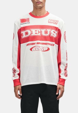 Persona que lleva una camisa de malla blanca y roja de manga larga con "DEUS" y logotipos personalizados de motocicletas, combinada con pantalones negros.
