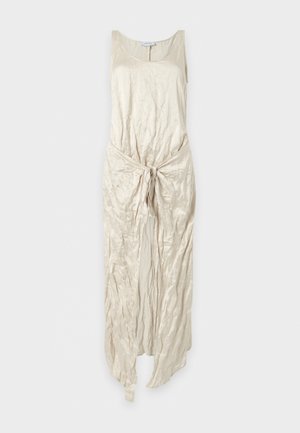 Femme aux longs cheveux foncés portant une robe crème sans manches, longueur genoux, avec un nœud à l'avant, debout pieds nus sur un fond blanc.